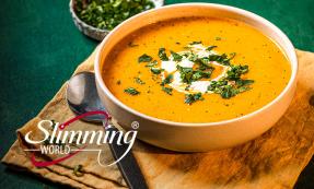 Carrot and coriander soup in a white bowl with herbs sprinkled on top. The bowl is on top of a brown cloth with a spoon to the left.