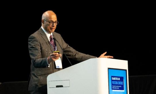 Photo of a man dressed in a grey suit gesturing and speaking on a podium