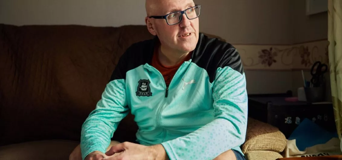 Paul sits in a light blue jacket on a sofa looking to his left